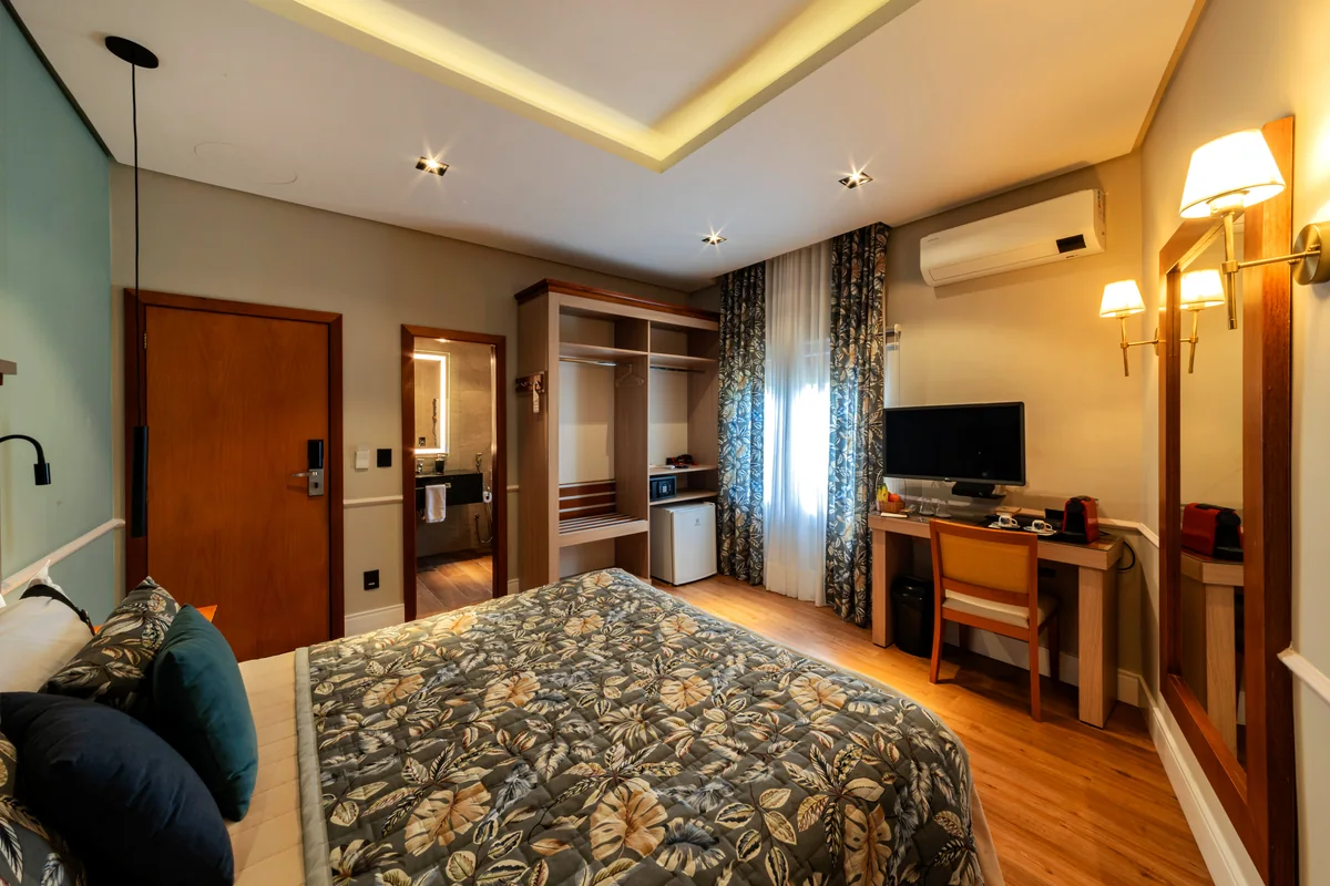Quarto Queen do Hotel Britânico, com cama de casal, closet aberto, escrivaninha com cafeteira e banheiro ao fundo
